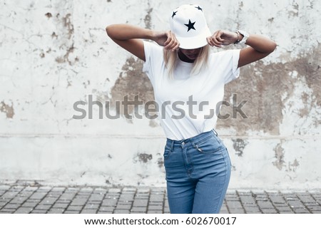 Hipster girl wearing blank white t-shirt, jeans and baseball cap posing against rough street wall, minimalist urban clothing style, mock up for tshirt print store
