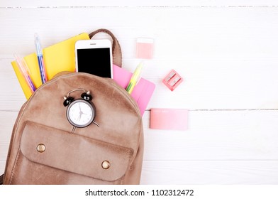 Hipster grey leather backpack, full of school supplies, blank screen cell phone, earphones, pink & yellow notebook, glasses, mini alarm clock. Back to school concept. Close up, copy space, flat lay. - Powered by Shutterstock