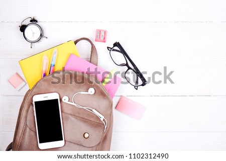 Hipster grey leather backpack, full of school supplies, blank screen cell phone, earphones, pink & yellow notebook, glasses, mini alarm clock. Back to school concept. Close up, copy space, flat lay.