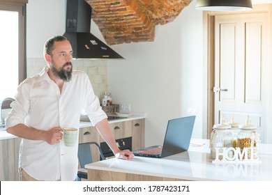 Hipster Guy With A Beard Drinking Coffee While Working On His Laptop At Home