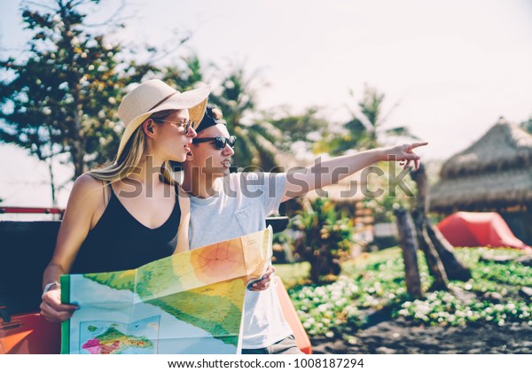Hipster guy pointing on road in tropical environment
talking with her girlfriend traveling with during summer
vacation,romantic couple searching right direction on map for
driving on rental car