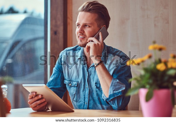 Hipster guy with a stylish haircut and beard\
sits at a table in a roadside cafe, talking on the phone and holds\
a tablet computer.
