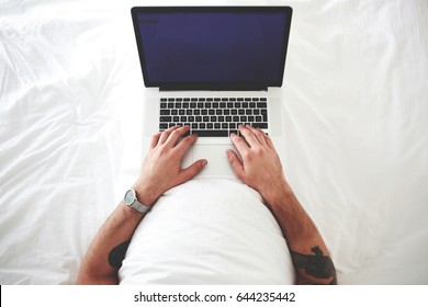 Hipster Guy With Tatoo On Hands Is Booking O-nline Ticket On A Plane Via Laptop Computer, While Is Lying On A Bed In Hotel.Man Is Working On Net-book With Blank Copy Space Screen For Advertise Content