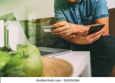 hipster hand using smart phone and laptop computer,holding credit card payments online business,sitting on sofa in living room,green apples in wooden tray,filter