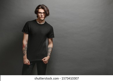 Hipster Handsome Male Model With Glasses Wearing Black Blank T-shirt And Black Jeans With Space For Your Logo Or Design In Casual Urban Style