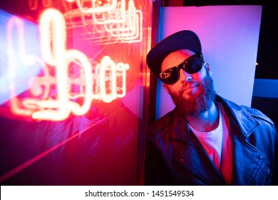 Hipster handsome man on the city streets being illuminated by neon signs. He is wearing leather biker jacket or asymmetric zip jacket with black cap, jeans and sunglasses.