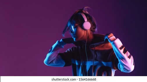 Hipster igen teen pretty fashion girl model wear stylish glasses headphones enjoy listen new cool music mix stand at purple studio background in trendy 80s 90s club blue party light, profile view