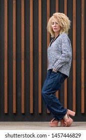 Hipster Image. European Girl Blonde Hairstyle Afro Curls Originally Posing In Full Growth In Gray Knitted Sweater, Free Blue Jeans And Chunky Hell.