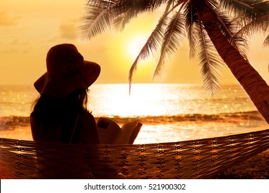  
Hipster Lady With Sunhat Reading Book In Hammock Under Coconut Palm Tree At Tropical Beach With Sunset And Water Bokeh Background. Woman Silhouette At The Beach, Travel Destination.
  