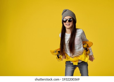hipster little child girl in hat and sunglasses with yellow skateboard looking super excited on yellow background. copy space