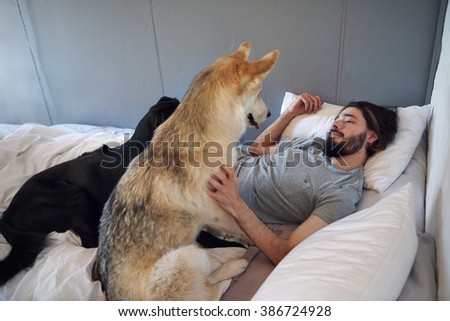 hipster man with beard looks lovingly at pet dogs lying in his bed while he wakes up in the morning