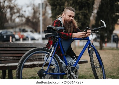Hipster man with a beard using a smart phone on a bench outdoors, mountain bike propped against the bench in an urban park.