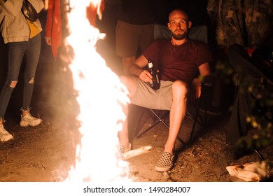 Hipster Man Drinking Beer And Relaxing With Friends Travelers At Big Bonfire At Night Camp In The Forest. Group Of People Resting Near Fire In The Evening, Camping Near Lake