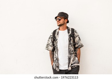 Hipster Man In Fashion Summer Clothes With Backpack Stands Near White Wall. Guy Travel On The Street