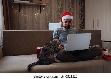 A Hipster Man With Grey Cat Sitting On A Sofa At Home At Christmas Time. Virtual Christmas House Party. Online Team Meeting Video Conference Calling From Home. Man Wearing Santa Hat