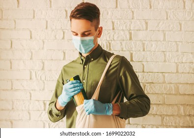 Hipster Man In Medical Gloves And Protective Mask On Face On Shopping In Grocery Store. Young Man Shopper Put Spaghetti In Cotton Eco Tote Bag. Personal Protection For Customer, Coronavirus COVID-19