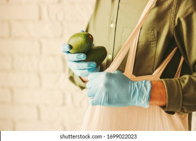 Hipster Man Shopper Putting Fresh Ripe Avocado In Cotton Eco Tote Bag In Supermarket. Young Man In Protective Medical Gloves Shopping Healthy Food In Store. Personal Safety, COVID-19, Coronavirus