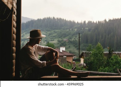 hipster man sitting on porch of wooden house  looking at mountains in evening, calm moment, summer vacation concept, space for text