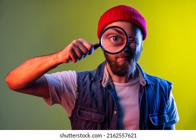 Hipster man standing, holding magnifying glass and looking at camera with big zoom eye, funny expression, wearing hat and denim vest. Indoor studio shot isolated on colorful neon light background.