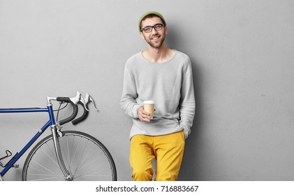 Hipster Man Standing With Takeaway Coffee, Smiling Plesantly Into Camera, Going To Have Trip With His Friends On Bicycle. Happy Carefree Handsome Guy In Eyewear Isolated Over Grey Background