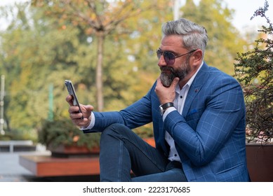 A Hipster Mature Business Man Making A Video Call From His Mobile Phone While Sitting On The Park Bench.	