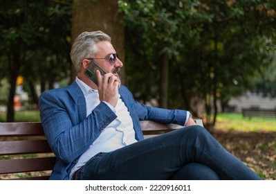 A Hipster Mature Business Man Talking On Mobile Phone While Siting At The Park Bench.