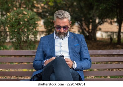 A Hipster Mature Business Man Using A Mobile Phone App Texting While Sitting On The Park Bench.