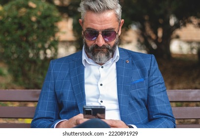 A Hipster Mature Business Man Using A Mobile Phone App Texting While Sitting On The Park Bench.