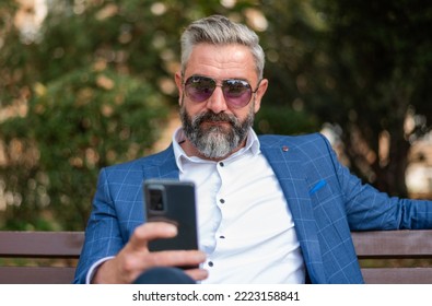 A Hipster Mature Business Man Using A Mobile Phone App Texting Outside While Sitting On The Park Bench.