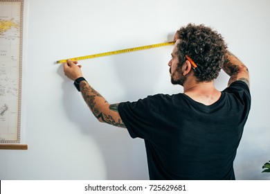 Hipster Millennial Man With Arm Tattoos And Curly Hair, Small Business Owner, Contractor Measures Wall To Start Reconstruction Or Adapt For Painting, New Home Ownership 