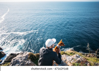 Hipster millennial young man in hoodie sweatshirt and five panel cap sits on cliff and overlooks sunset over ocean together with best friend, basenji dog