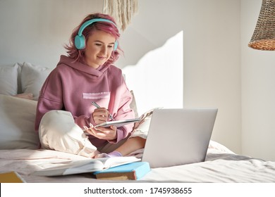 Hipster teen girl school student with pink hair wear headphone write notes watch video online webinar learn on laptop sit in bed distance elearning course video conference pc call in bedroom at home. - Shutterstock ID 1745938655
