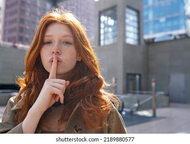 Hipster teen stylish cool redhead fashion girl model showing shh sign asking to keep secret, be hush silent or privacy silence standing in big city urban location. Headshot portrait