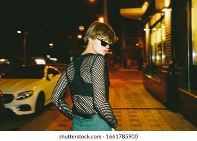Hipster Teen Tomboy Girl Short Hairstyle Wear Stylish Glasses Clothes Walking On Late Night Urban Dark City Street Look At Camera. Fashion Trendy Stylish Teenager Young Woman 80s 90s Style Outdoors.