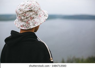 Hipster Teenager In Bucket Hat Standing On Top Of Rock Mountain And Looking On River, View From Back. Atmospheric Calm Moment. Wanderlust And Travel. Copy Space