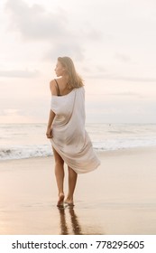 Hipster Trendy Girl In Casual Summer Dress Walking Barefoot By The Waterline And Look To The Waves. Sporty Lady On Sea Sand Beach Sunset Or Ocean Sunrise. Travel, Active, Yoga Lifestyle Concept.