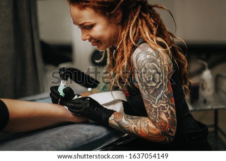Hipster woman girl with tattoos and dreadlocks makes a tattoo on the female leg, concept by tattoo artist and creative work