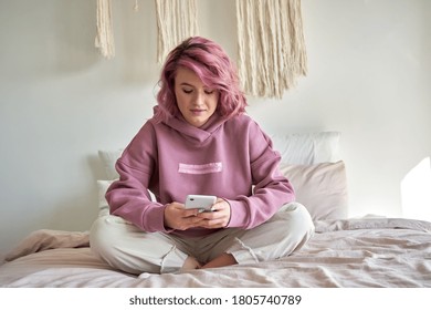 Hipster woman with pink hair holding smart phone chatting in social media, watching video content, texting messages, playing games, using mobile apps, surfing internet sit in bed at home. Vertical - Powered by Shutterstock