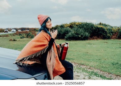 Hipster Woman sits on car's hood, wrapped in warm Shawl Wraps Poncho Cape and hat and enjoying warm drink, while overlooking rural landscape during road trip. Cold season travel, vacation concept - Powered by Shutterstock