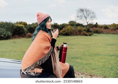 Hipster Woman sits on car's hood, wrapped in warm Shawl Wraps Poncho Cape and hat and enjoying warm drink, while overlooking rural landscape during road trip. Cold season travel, vacation concept - Powered by Shutterstock