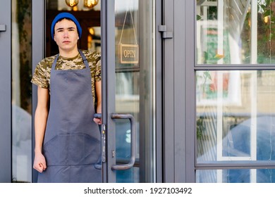 Hipster Young Handsome Man Standing On Doorway Near Glass Window. Portrait Of Sad Serious Young Worker Putting Open Chalkboard On Cafe Door.