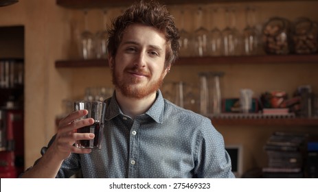 Hipster Young Man Smiling And Drinking A Glass Of Coke With Bar Wooden Shelves On Background