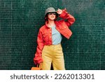 Hipster young woman in bright clothes, sun glasses, backpack bag and bucket hat posing on the green tile wall background. Urban city street fashion. Fashion blogger. Selective focus. Copy space