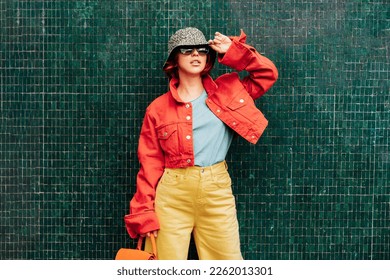 Hipster young woman in bright clothes, sun glasses, backpack bag and bucket hat posing on the green tile wall background. Urban city street fashion. Fashion blogger. Selective focus. Copy space - Powered by Shutterstock