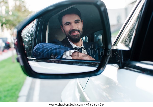 His smile in a mirror. Handsome young businessman\
looking at camera at mirror with smile\
while sitting on front\
passenger seat\
