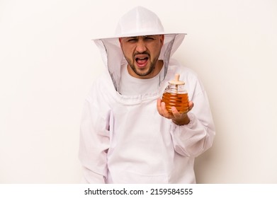 Hispanic Beekeeper Man Holding Honey Isolated On White Background Screaming Very Angry And Aggressive.