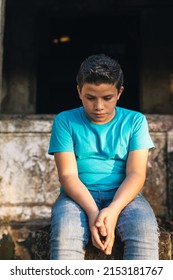 Hispanic Boy Sitting On Some Ruins Looking At The Ground, With A Sad Expression.