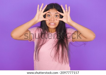 Hispanic brunette girl wearing pink t-shirt over purple background keeping eyes opened to find a success opportunity.