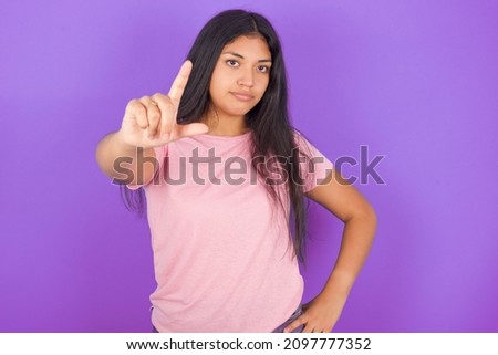 Hispanic brunette girl wearing pink t-shirt over purple background making fun of people with fingers on forehead doing loser gesture mocking and insulting.