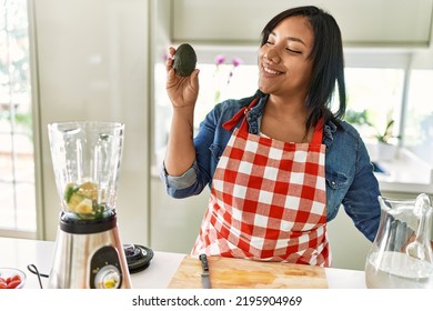 Hispanic Brunette Woman Preparing Fruit Smoothie Holding Avocado At The Kitchen